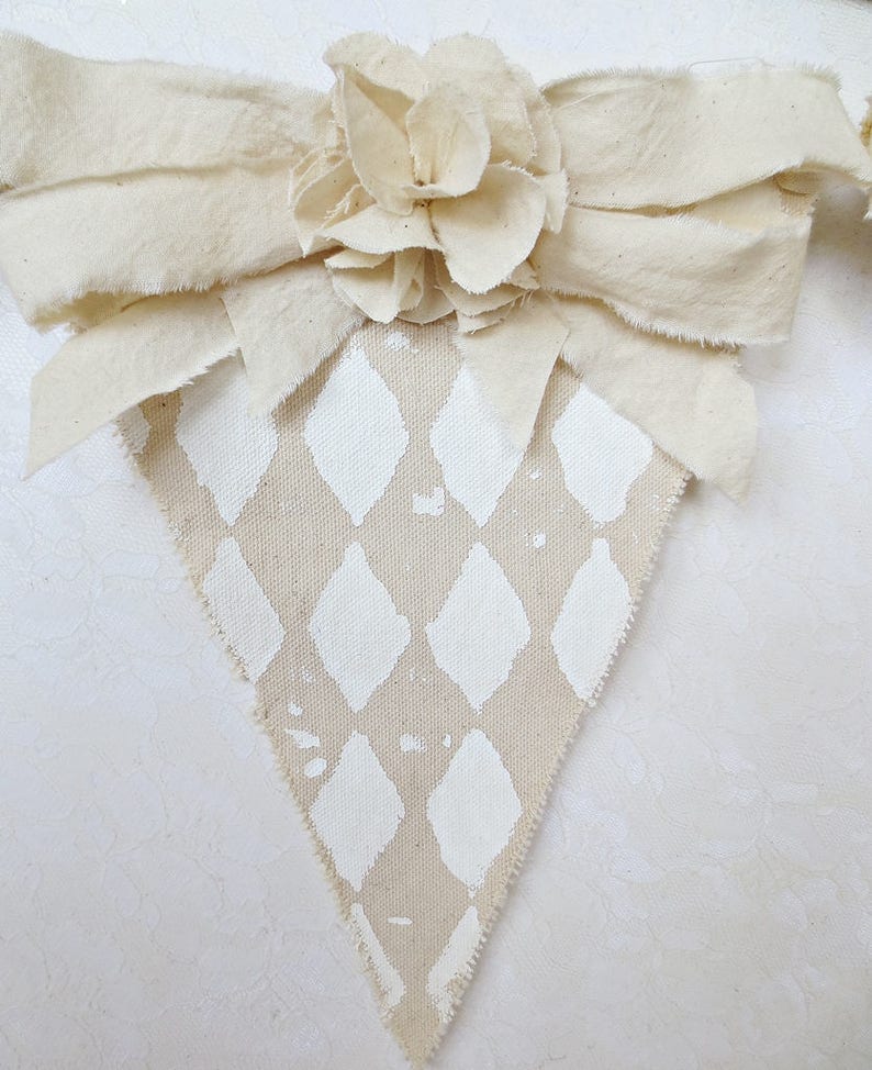 May include: A beige fabric triangle banner with a white diamond pattern. The banner is decorated with a large white fabric flower.
