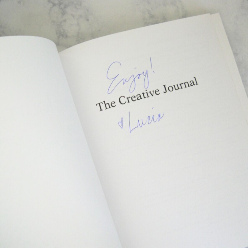 Peut inclure: Un livre blanc avec les mots "Enjoy! The Creative Journal" &eacute;crits &agrave; l'encre bleue. Sous le texte se trouve un symbole de c&oelig;ur et le nom "Lucia" &eacute;crit &agrave; l'encre bleue.