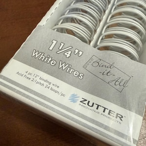 May include: A package of Zutter Bind-it-All 1 1/4 inch white binding wires. The packaging contains 4 pieces of 12 inch acid-free binding wire with a 2:1 pitch and 24 loops per piece. The white metal wire coils are visible through the clear plastic wrap.