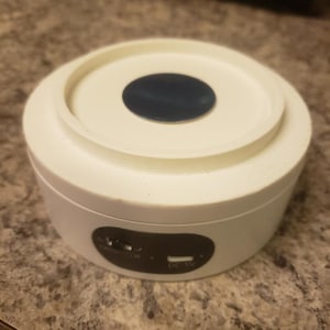 May include: A white, round electronic device with a dark blue center and a black panel with ports. The device has a circular top and a cylindrical base. The surface is smooth and the overall design is minimalist.