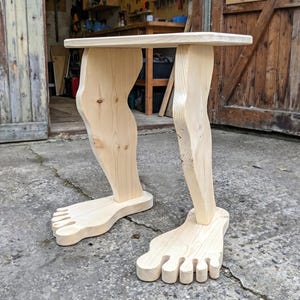Può includere: Un tavolo in legno stravagante con un piano rettangolare sostenuto da due gambe scolpite a forma di gambe e piedi umani. Il tavolo è realizzato in legno chiaro e presenta un design artistico unico. L'altezza del tavolo è di circa 60 cm.