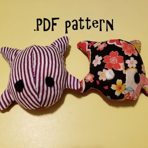 May include: Two handmade plush toys in the shape of fish. One fish is purple and white striped with black eyes. The other fish is black with a floral pattern and white eyes. The text '.PDF pattern' is written above the toys.