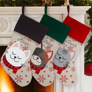 May include: Three personalized Christmas stockings with a cat theme. The stockings are red, green, and black with a white and brown burlap fabric. The stockings are decorated with snowflakes and stars. The names "Alan", "Kirby", and "Mittens" are written on the stockings.