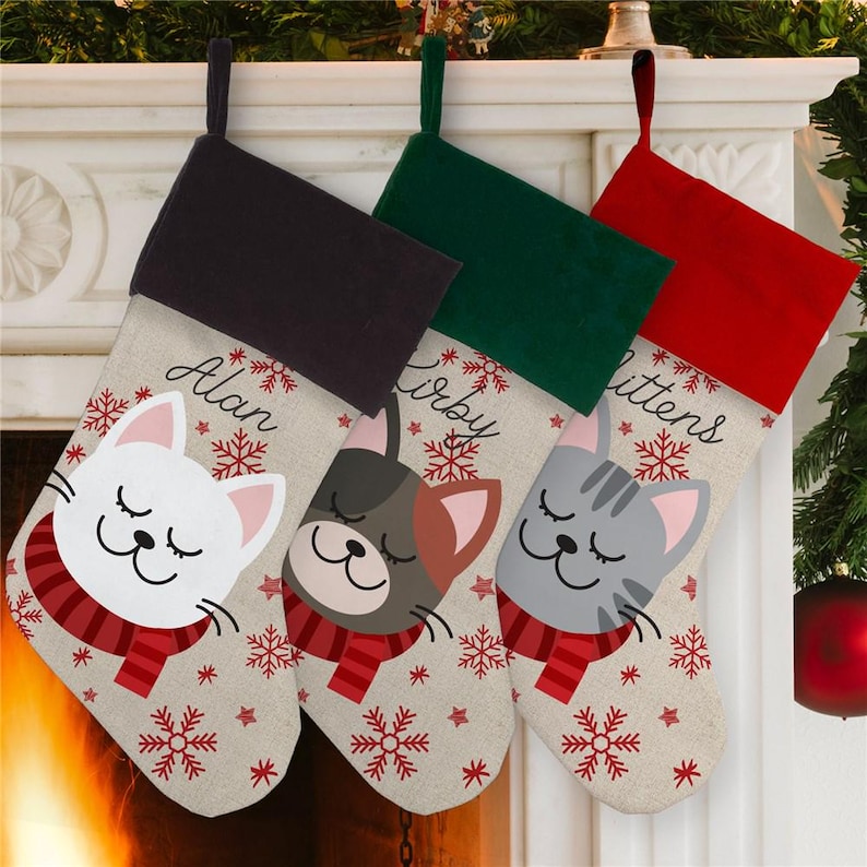 May include: Three personalized Christmas stockings with a cat theme. The stockings are red, green, and black with a white and brown burlap fabric. The stockings are decorated with snowflakes and stars. The names "Alan", "Kirby", and "Mittens" are written on the stockings.