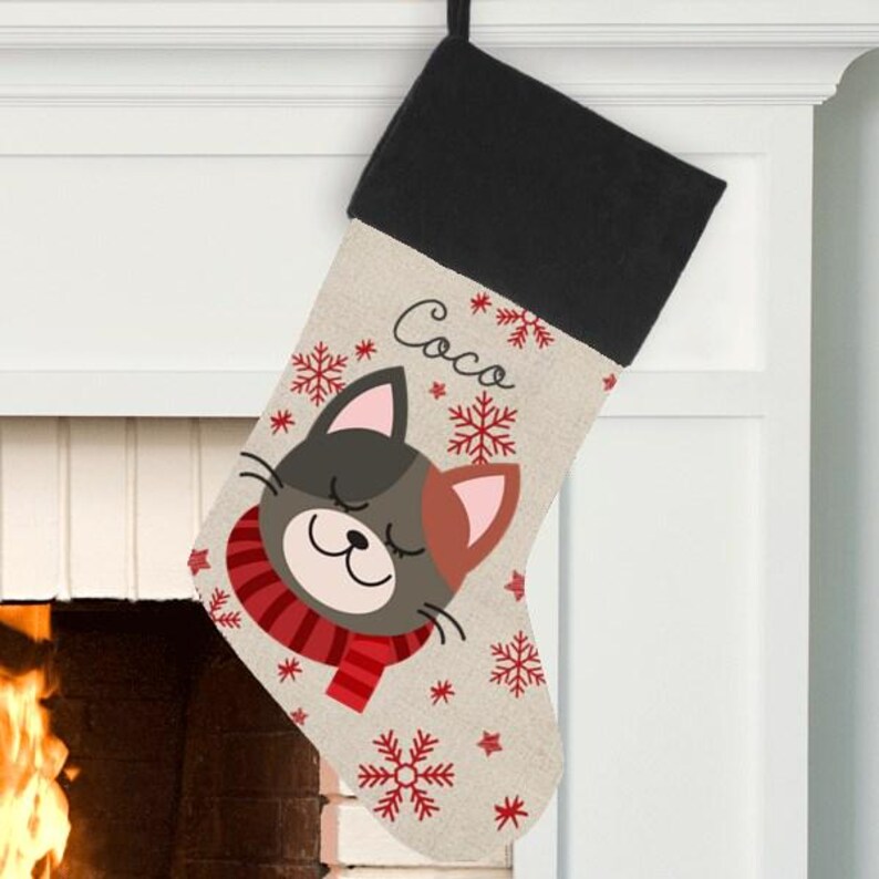 May include: Personalized Christmas stocking with a cute cartoon cat wearing a red scarf and snowflakes. The stocking is beige with a black cuff and the name "Coco" is printed on the front.