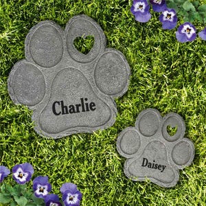May include: Two gray paw print stepping stones with heart-shaped cutouts. One stone reads "Charlie," the other "Daisey." The stones are set in green grass, with purple and white pansies scattered around.