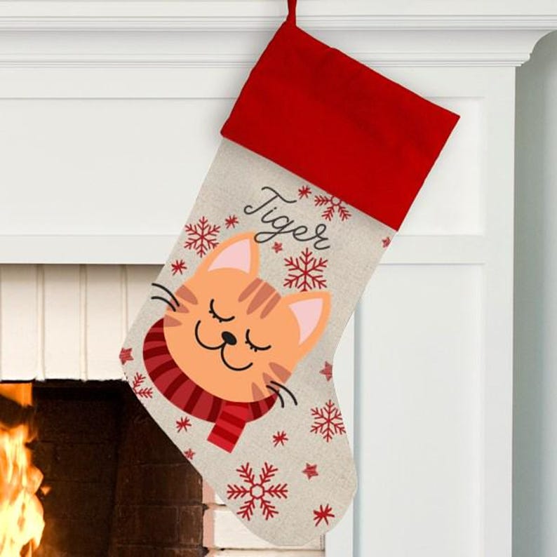 May include: Personalized Christmas stocking with a cute orange cat wearing a red scarf and snowflakes. The stocking is made of burlap and has the name "Tiger" written on it.