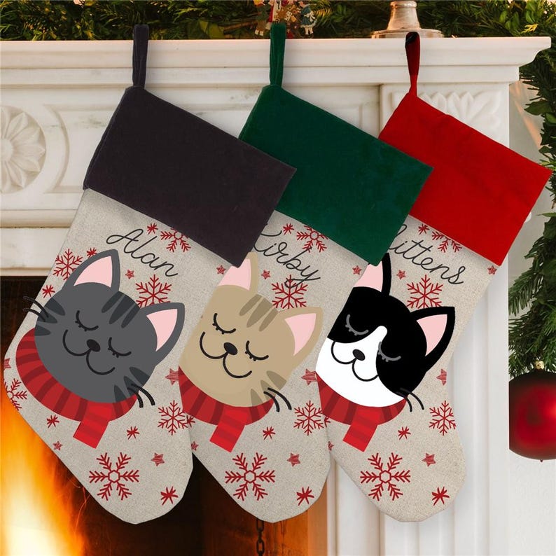 May include: Three personalized Christmas stockings with a cat design. The stockings are made of a beige fabric with red and white snowflakes. The top of each stocking is a different color: black, green, and red. The names "Alan", "Kirby", and "Mittens" are printed on the stockings.