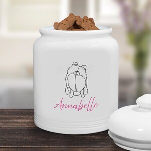 Może przedstawiać: Biały ceramiczny słoik na przysmaki dla ps&oacute;w z pokrywką. Słoik przedstawia czarny rysunek psa i imię "Annabelle" w r&oacute;żowym skrypcie. Słoik jest wypełniony psimi herbatnikami.