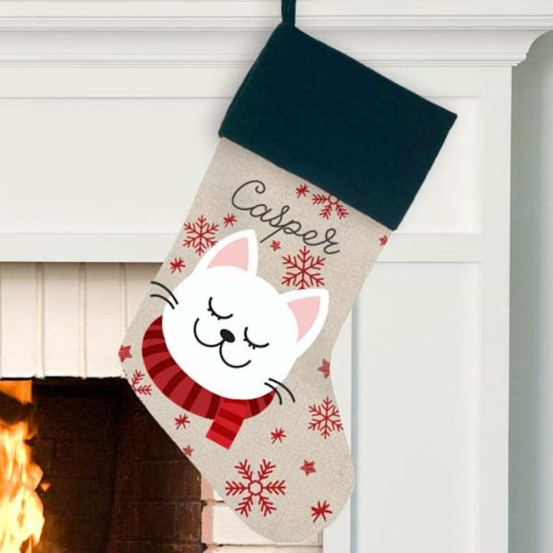 May include: Personalized Christmas stocking with a white cat wearing a red scarf and snowflakes. The stocking is beige with the name "Casper" embroidered on it.