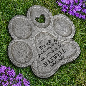 May include: A gray, paw-shaped garden stone with a heart-shaped cutout and the inscription "You left paw prints on our hearts MAXWELL 2006-2023." The stone is surrounded by green grass and purple flowers.