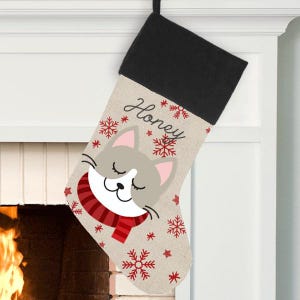 May include: Personalized Christmas stocking with a cute cartoon cat wearing a red scarf and snowflakes. The stocking is beige with a black cuff and the name "Honey" is embroidered on the front.