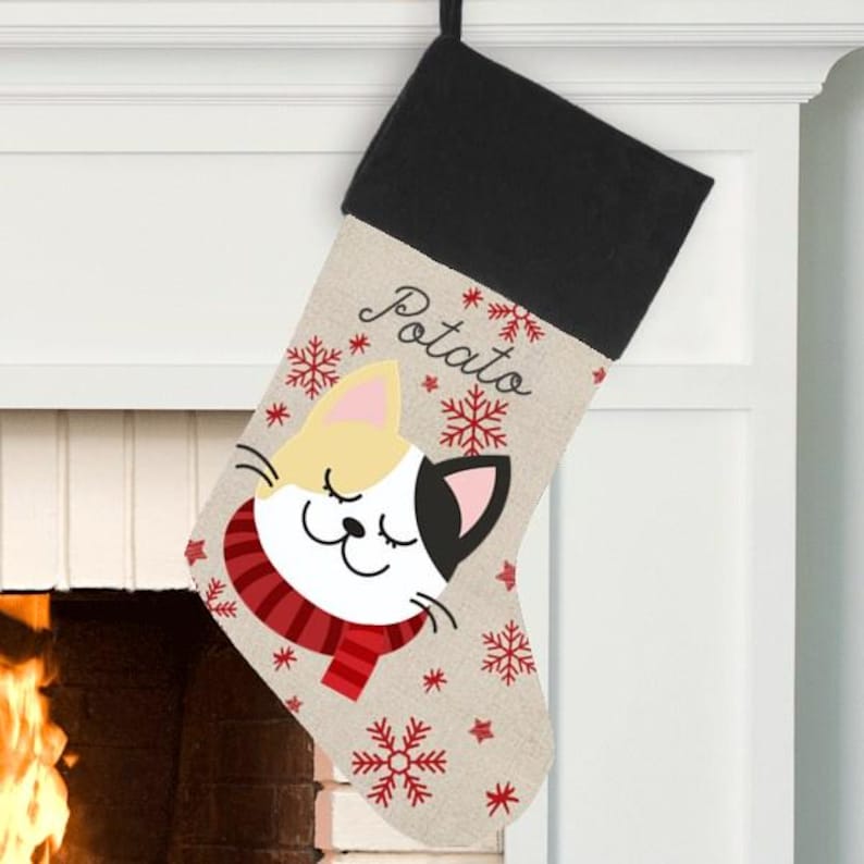 May include: A beige Christmas stocking with a black cuff, featuring a cartoon cat with a red scarf and snowflakes. The stocking is personalized with the name "Potato".