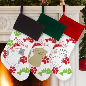 May include: Three Christmas stockings with black, green, and red tops. Each stocking features a cartoon cat wearing a Santa hat, with the names "Mittens", "Oliver", and "Loki". The stockings are decorated with paw prints and fish bones.