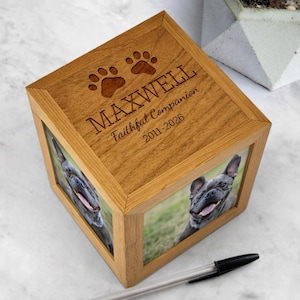 May include: A wooden pet memorial box featuring a dog's photo on two sides. The top is engraved with "MAXWELL", paw prints, and "Faithful Companion 2011-2026". The box is made of light-colored wood.