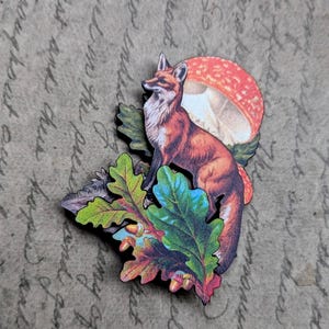 May include: A wooden fox figurine with a red and white mushroom and colorful oak leaves. The fox is orange and white, with a detailed design. The leaves are green, blue, and red. The background is a textured, tan surface.
