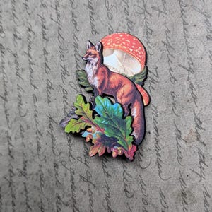 May include: A wooden fox and mushroom cut-out with colorful leaves. The fox is reddish-brown with a white chest, standing near a red and white mushroom. The leaves are green, blue, and orange.