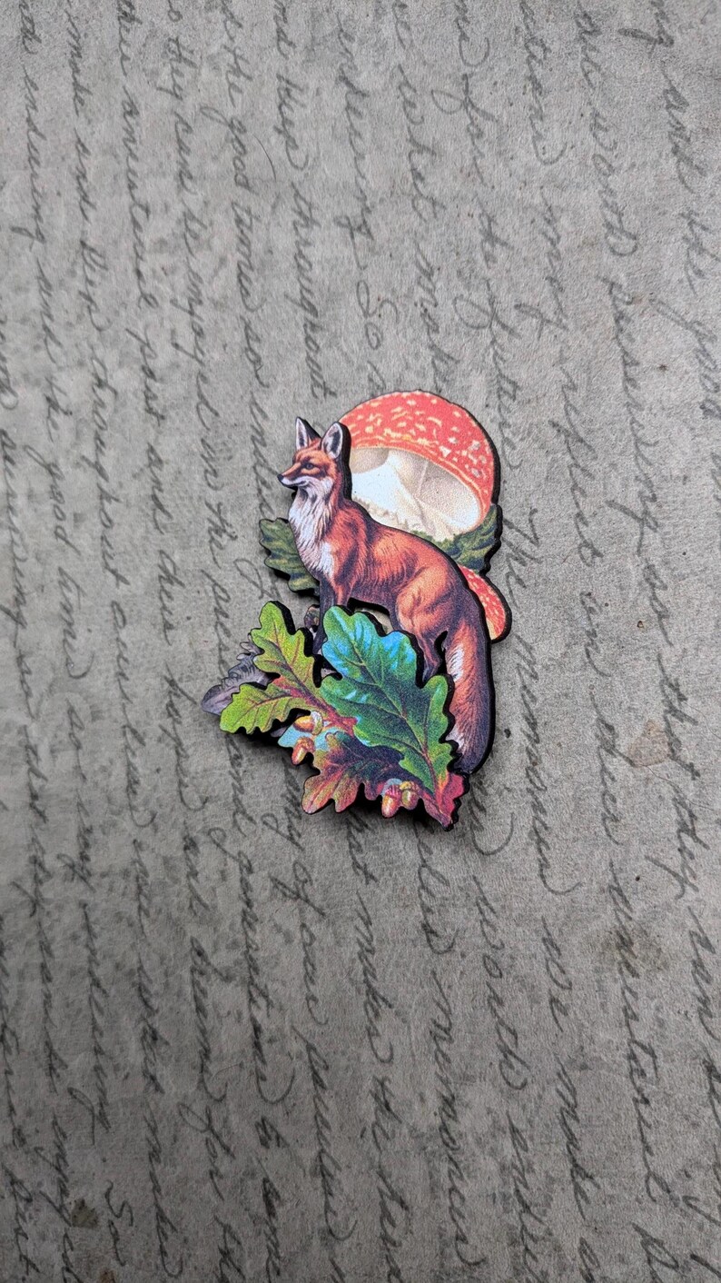 May include: A wooden fox and mushroom cut-out with colorful leaves. The fox is reddish-brown with a white chest, standing near a red and white mushroom. The leaves are green, blue, and orange.
