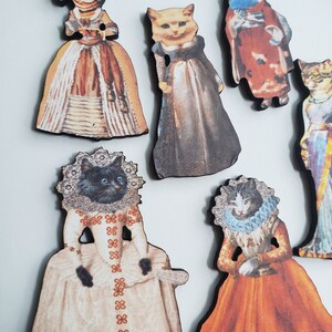 May include: A set of nine wooden ornaments featuring cats dressed in vintage clothing. The cats are wearing various colors and patterns, including red, white, blue, and black. The ornaments are perfect for adding a touch of whimsy to any home decor.