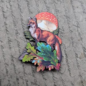 May include: A wooden fox figurine with a red and brown coat, standing on colorful leaves and a mushroom. The fox is facing left. The background is a vintage paper with handwritten text.