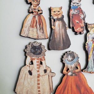 May include: Set of eight wooden ornaments featuring cats dressed in vintage clothing. The ornaments are painted with intricate details and feature a variety of colors, including brown, white, black, orange, and blue.