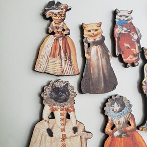 May include: Set of eight wooden cat figurines dressed in vintage clothing. Each figurine features a different cat breed with a unique outfit and pose. The figurines are painted in a variety of colors, including brown, white, black, and orange.
