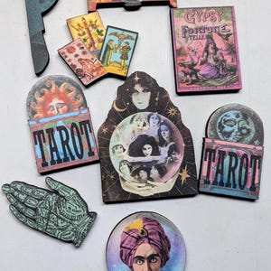 May include: A collection of tarot-themed wooden cutouts and cards. The designs include images of a fortune teller, a crystal ball, the sun, and the moon. The text "TAROT" is featured on two of the cutouts. The color palette includes shades of blue, pink, and gold.