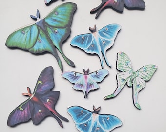 May include: A collection of colorful moth-shaped wooden cutouts. The moths feature various shades of blue, green, purple, and black, with intricate wing patterns. Some have a coin next to them for size reference. The cutouts are on a white surface.