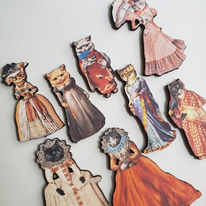 May include: Nine wooden ornaments featuring cats dressed in vintage clothing. The cats are wearing dresses, hats, and other accessories. The ornaments are painted in a variety of colors, including pink, red, blue, and green.