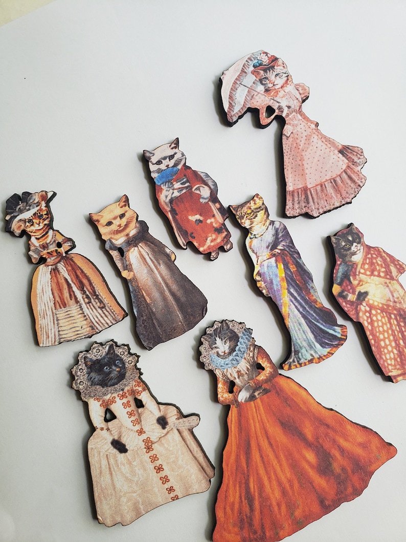 May include: Nine wooden ornaments featuring cats dressed in vintage clothing. The cats are wearing dresses, hats, and other accessories. The ornaments are painted in a variety of colors, including pink, red, blue, and green.