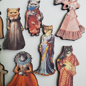 May include: A set of eight wooden cat figurines dressed in various vintage clothing. The cats are wearing dresses, suits, and a sari. Some of the cats are holding umbrellas and other accessories.