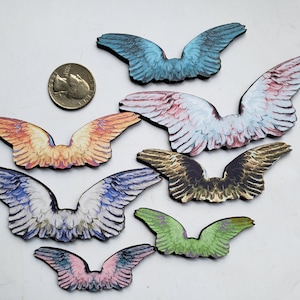 May include: A collection of seven wooden wing cutouts in various colors, including blue, pink, green, and orange. Each wing has a detailed, feathered design. A quarter is included for size reference.