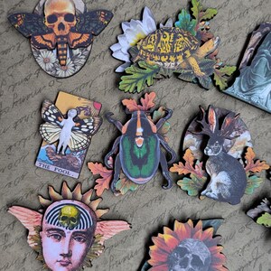 May include: A collection of decorative wooden cutouts featuring various designs. Includes a skull moth, a turtle with flowers, a statue, a tarot card, a beetle, a rabbit, a fox, a face with a skull, and a sunflower with a skull. The cutouts are on a textured background.