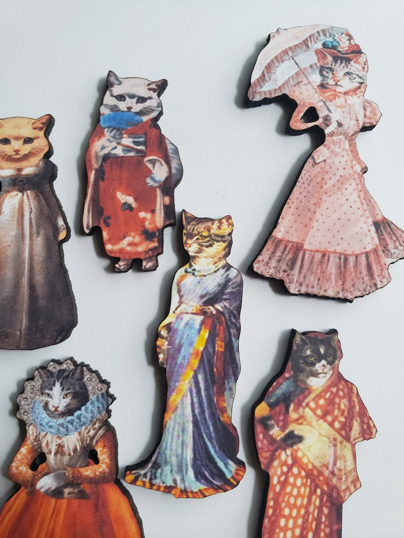 May include: Six wooden cat figurines dressed in different outfits. The cats are wearing a variety of clothing, including a kimono, a sari, and a Victorian dress. The figurines are all hand-painted and have a vintage look.