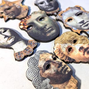 May include: A collection of flat, shaped wooden buttons featuring printed illustrations of classical human faces and busts. The designs include various expressions, hairstyles, and decorative collars in shades of gold, brown, grey, and blue.