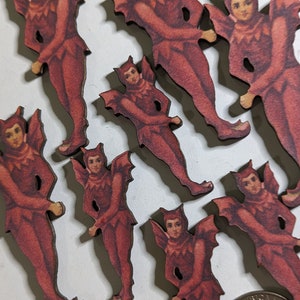 May include: A collection of wooden cutouts depicting figures in red devil costumes with bat wings. The figures have detailed facial features and are arranged on a white surface. A US quarter is included for size reference.