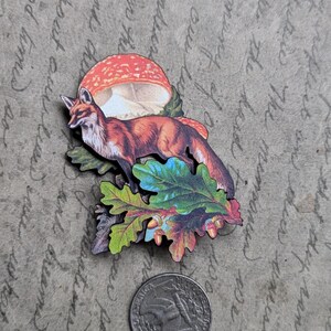 May include: A wooden fox and mushroom design with colorful leaves and acorns. The fox is orange and white, and the mushroom is red and white. A quarter is next to the design for scale.