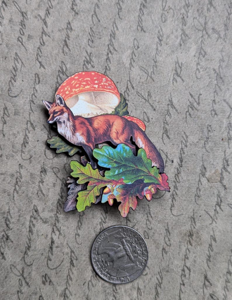 May include: A wooden fox and mushroom design with colorful leaves and acorns. The fox is orange and white, and the mushroom is red and white. A quarter is next to the design for scale.