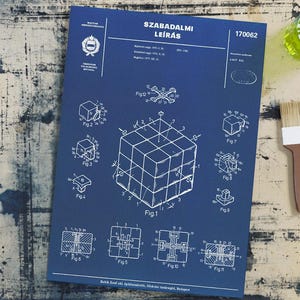 May include: A blue print with a diagram of a Rubik's Cube, labelled with figures and the text "SZABADALMI LEÍRÁS". A paint brush and a small jar with green liquid are on a distressed wooden surface next to the print.
