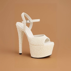 May include: White platform high heel sandal covered in small, round, pearl-like beads. The sandal has a thin ankle strap with a buckle, a peep-toe design, and a stiletto heel. The heel height is approximately 6 inches.