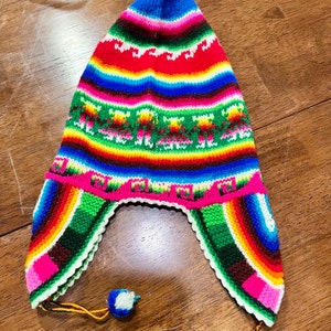 May include: A colorful, knitted hat with ear flaps. The hat features a vibrant rainbow pattern with stripes of red, green, yellow, blue, and pink. A small blue and white pom-pom hangs from the ear flap. The hat is a traditional style.