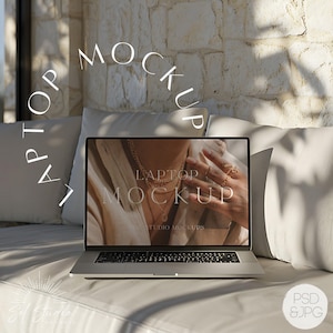 May include: A silver laptop computer with a white background on the screen. The screen displays the text "LAPTOP MOCKUP" in black. The laptop is sitting on a white couch with a white pillow behind it.