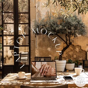 May include: A laptop computer with a white background on the screen that says "Laptop Mockup" is sitting on a wooden table with a cup of coffee and a smartphone. The laptop is in a cafe setting with a tree and a wall in the background.