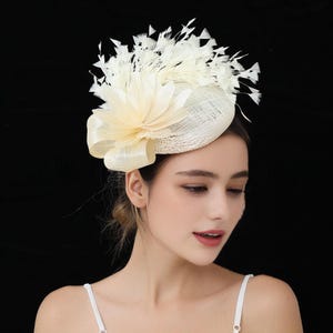 May include: A cream-colored fascinator with a woven base, adorned with a large, layered flower and a bow. White feathers fan out from the top, creating a dramatic effect. The headpiece is designed for special occasions.