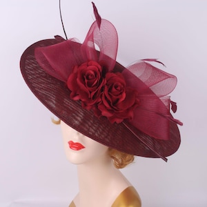 Peut inclure: Chapeau de cérémonie bordeaux à large bord, orné de deux grandes roses rouges en tissu et d'accents bordeaux transparents. Le chapeau présente un nœud décoratif et des plumes, idéal pour les occasions spéciales.