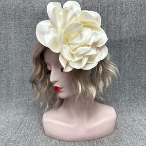 Peut inclure: Un accessoire pour cheveux en forme de fleur surdimensionnée de couleur crème. Les grands pétales superposés créent un design floral volumineux. L'accessoire est fixé sur une tête de mannequin avec des cheveux blonds et du rouge à lèvres rouge.