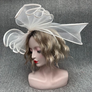 Pode incluir: Um elegante adorno de cabeça branco com um grande design de laço. O laço é feito de um tecido transparente e texturizado e é preso a uma tiara. O adorno de cabeça é exibido em uma cabeça de manequim com cabelo loiro e batom vermelho.