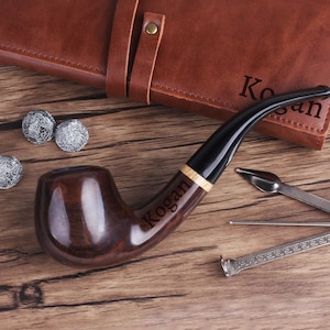 Peut inclure: Une pipe en bois brun avec un embout noir et une bande dorée. La pipe est gravée du nom "Kogan". La pipe est accompagnée d'un étui en cuir, d'un tamper et d'un nettoyeur de pipe.