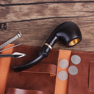 Może przedstawiać: Brązowy skórzany futerał na fajkę z czarną fajką, narzędziem do fajki i paczką siatek do fajki. Futerał ma zamek błyskawiczny i kieszeń na narzędzie do fajki.
