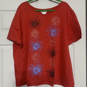 May include: Red short-sleeved t-shirt with a festive fireworks design in blue, black, and gold. The shirt has a crew neck and a relaxed fit.